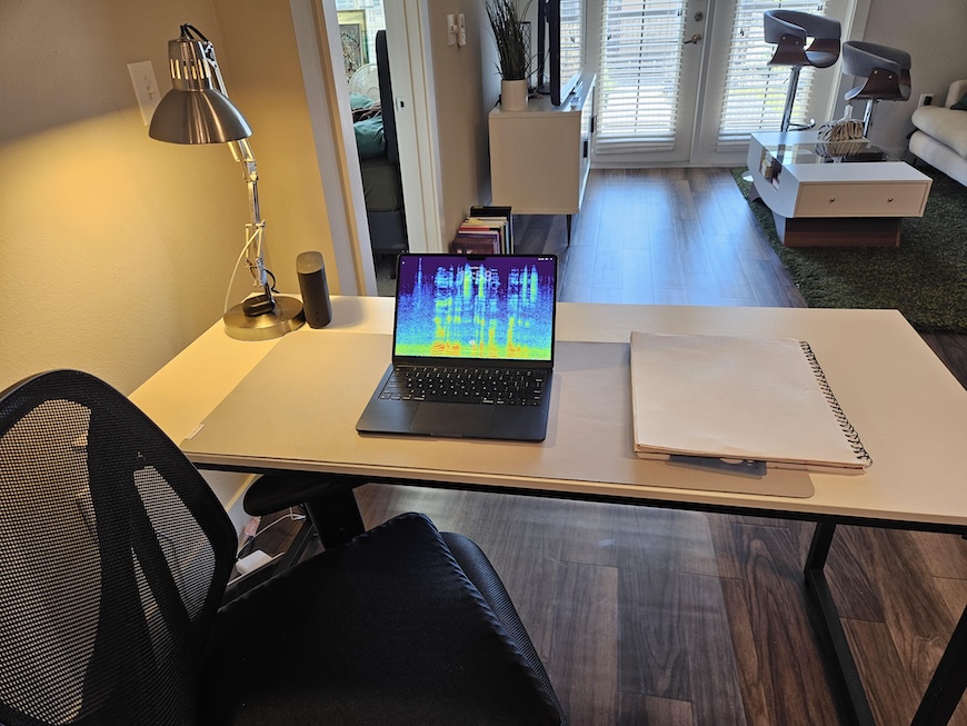 A simple desk setup with a MacBook Air, a spiral notepad, and a desk lamp. The laptop screen shows a colorful spectrogram. A mesh office chair is pulled up to a white desk in a living room.